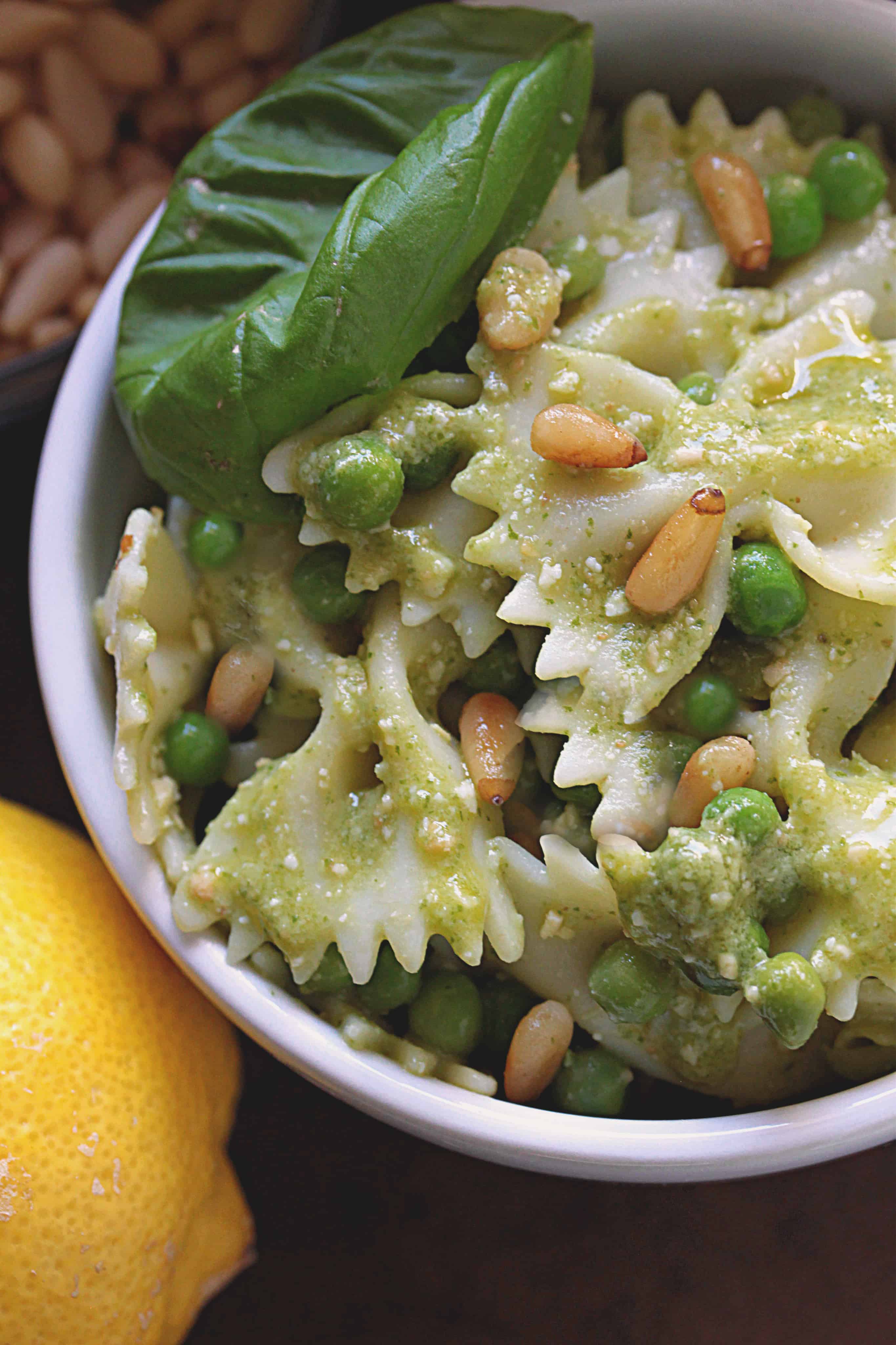 Bow Tie Pesto Pasta Salad with Peas in a white bowl garnished with peas, basil and pine nuts, with a lemon in the background