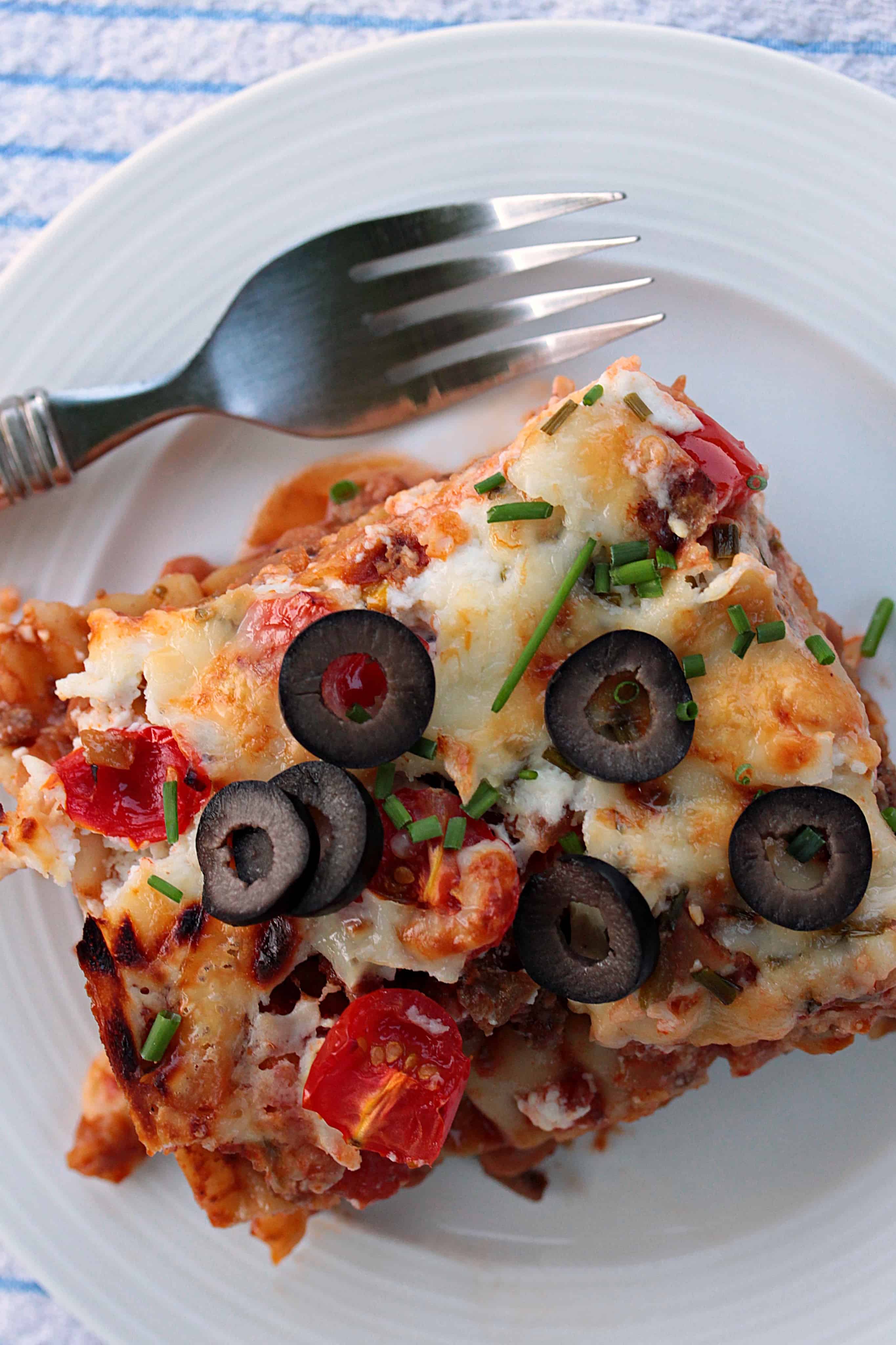 overhead photo of a piece of Mexican lasagna on a white plate topped with olives and green onion with a fork