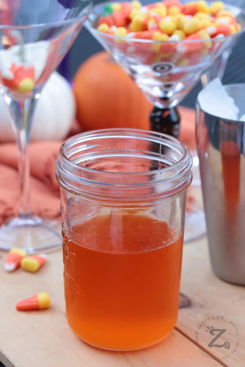 Candy Corn Vodka in a jar with a martini shaker, candy corn and pumpkins in the background