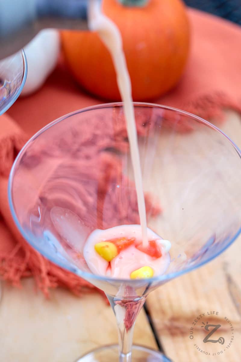 Candy Corn Martini being poured from a martini shaker into a martini glass with Candy Corn garnish and pumpkins in the background