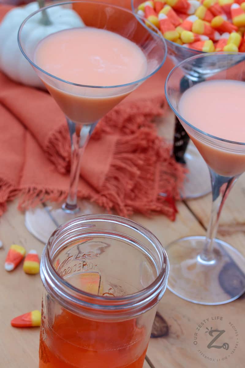 Two Candy Corn Martinis with Candy Corn Vodka in a jar and candy corns and pumpkin in the background