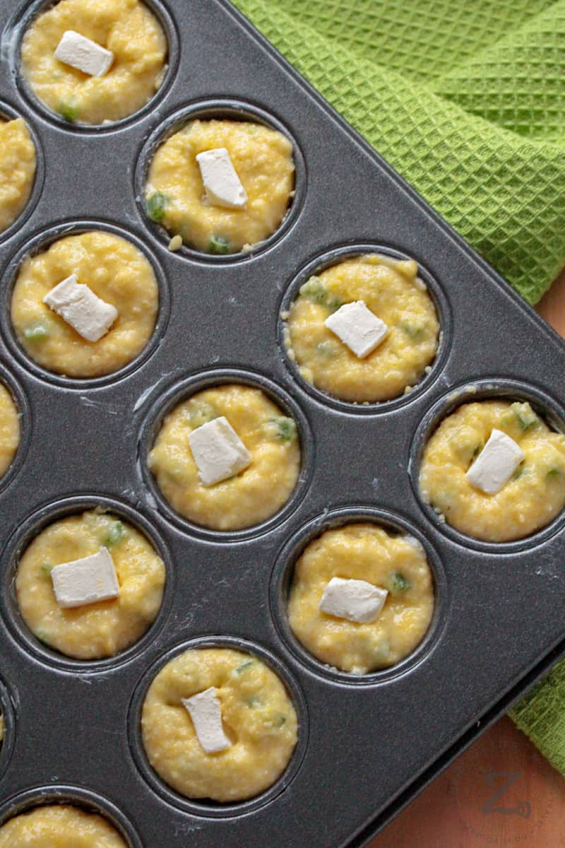 Mini Jalapeno Popper Corn Muffins with a cream cheese center in a buttered muffin tin with a green towel on the side