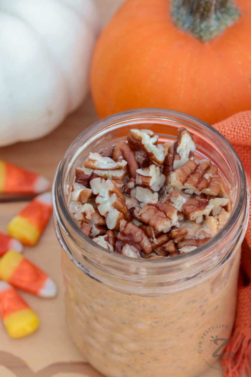 closeup of a mason jar filled with pumpkin overnight oats topped with pecans, with candy corn and an orange pumpkin and a white pumpkin in the background