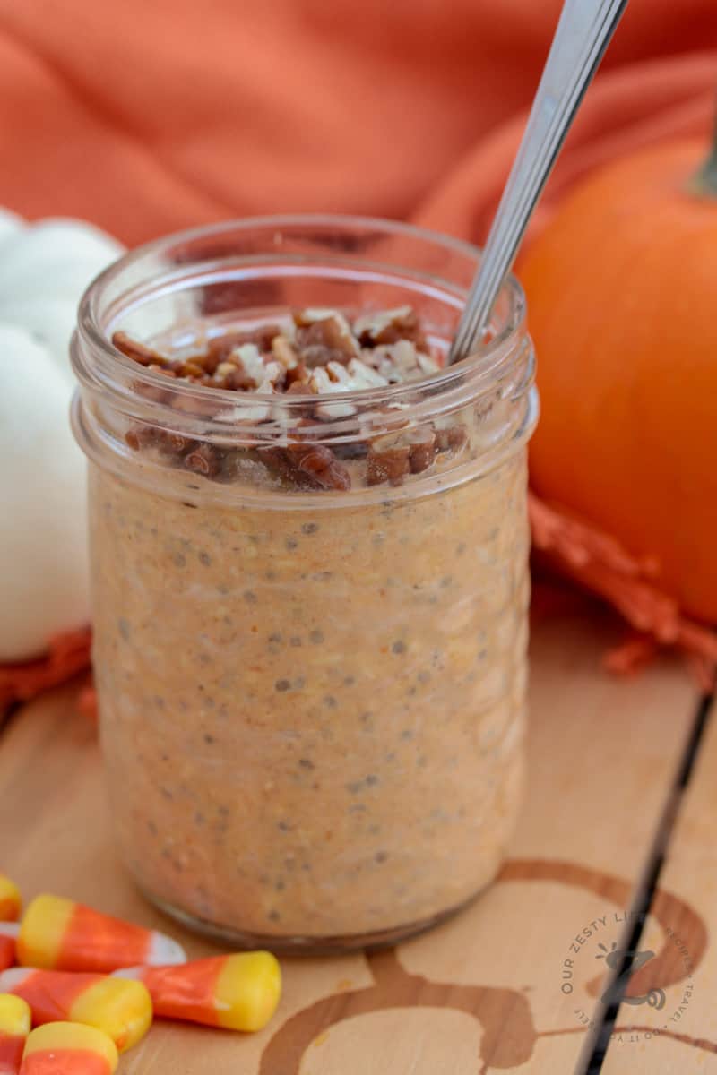 closeup of a mason jar filled with pumpkin overnight oats topped with pecans with a spoon in it, with candy corn and an orange pumpkin and a white pumpkin in the background