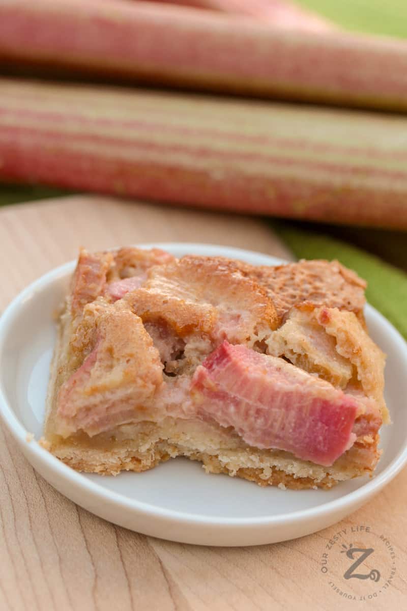 Closeup of shortbread rhubarb bars on a white plate with rhubarb stalks in the background