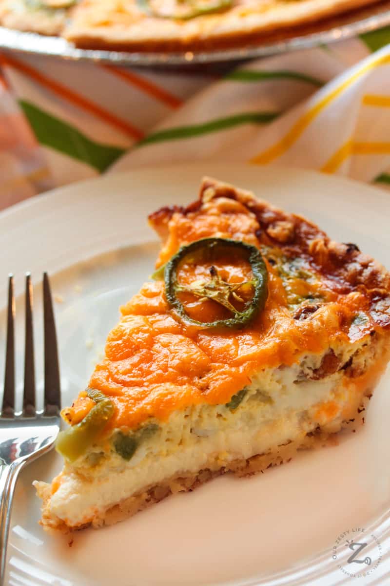 side view of a slice of Jalapeno Popper Quiche on a plate with a fork, with a pie tin on the sid