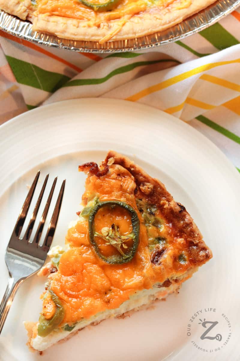 overhead of a slice of Jalapeno, Bacon and Cheese Quiche on a plate with a fork, with a pie tin on the side
