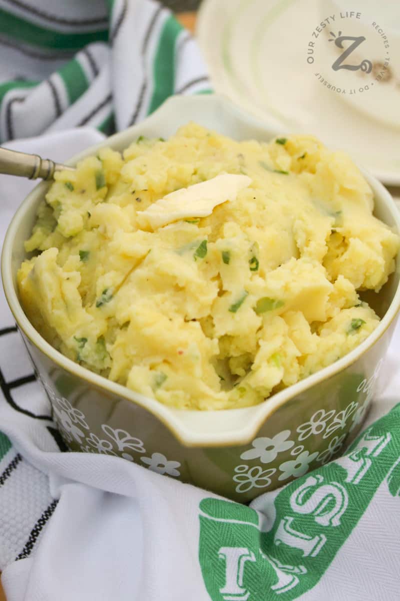 Irish Potato Recipe in a dish topped with a pat of butter with a spoon in it and Irish teatowels and plates in the background