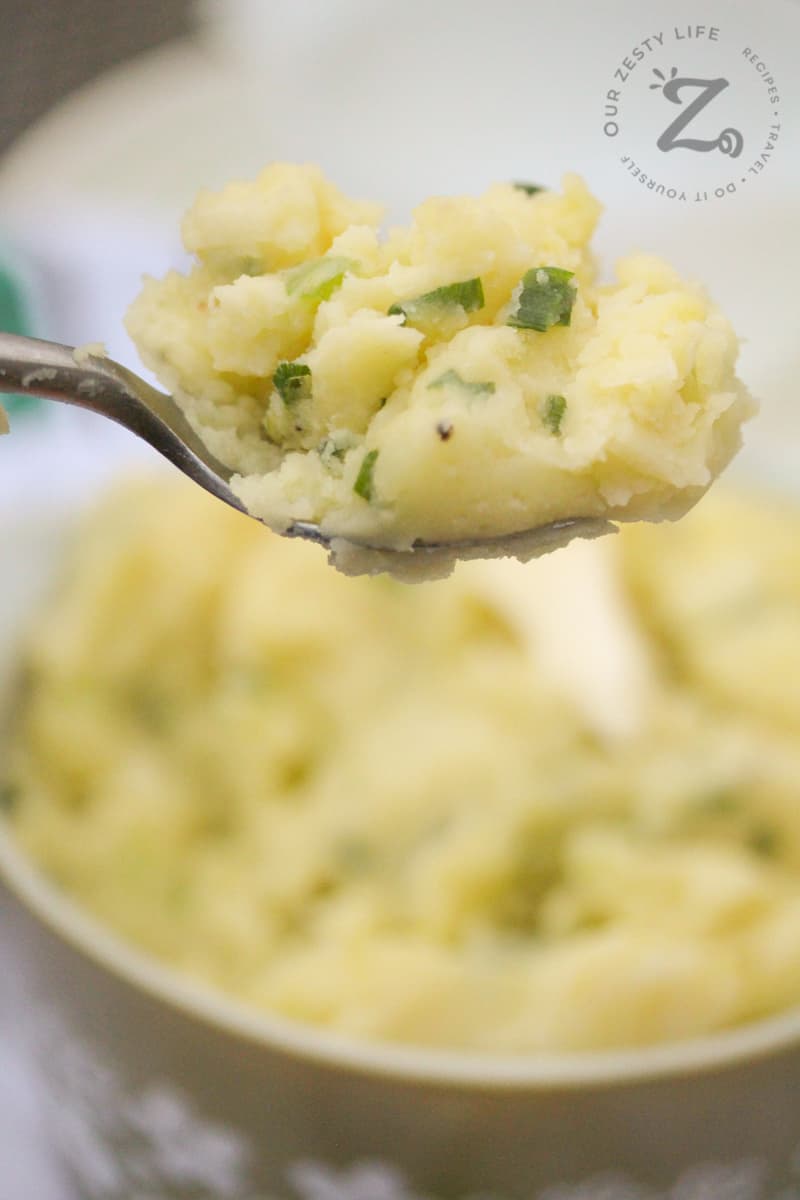 closeup of a bite of Irish Champ on a spoon with a serving dish of Irish mashed potatoes in the background