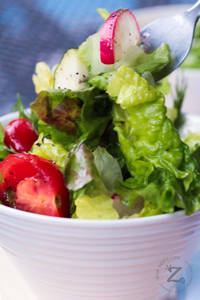 closeup of a forkful of red leaf salad with lemon vinaigrette, with fresh dill, radishes, grape tomatoes and peppers, with red leaf salad in a white bowl