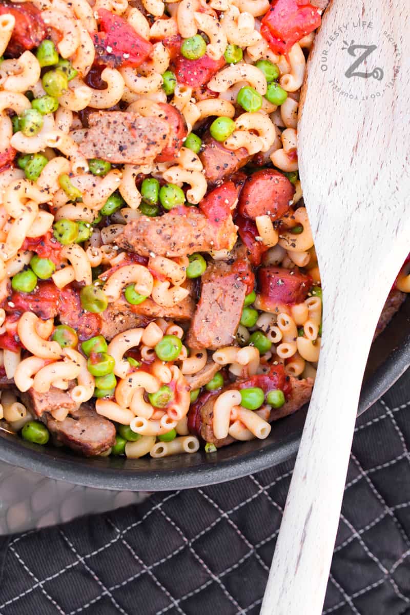 Overhead of hamburger pasta with sausage, peas and diced tomatoes in a skillet with a wooden spoon