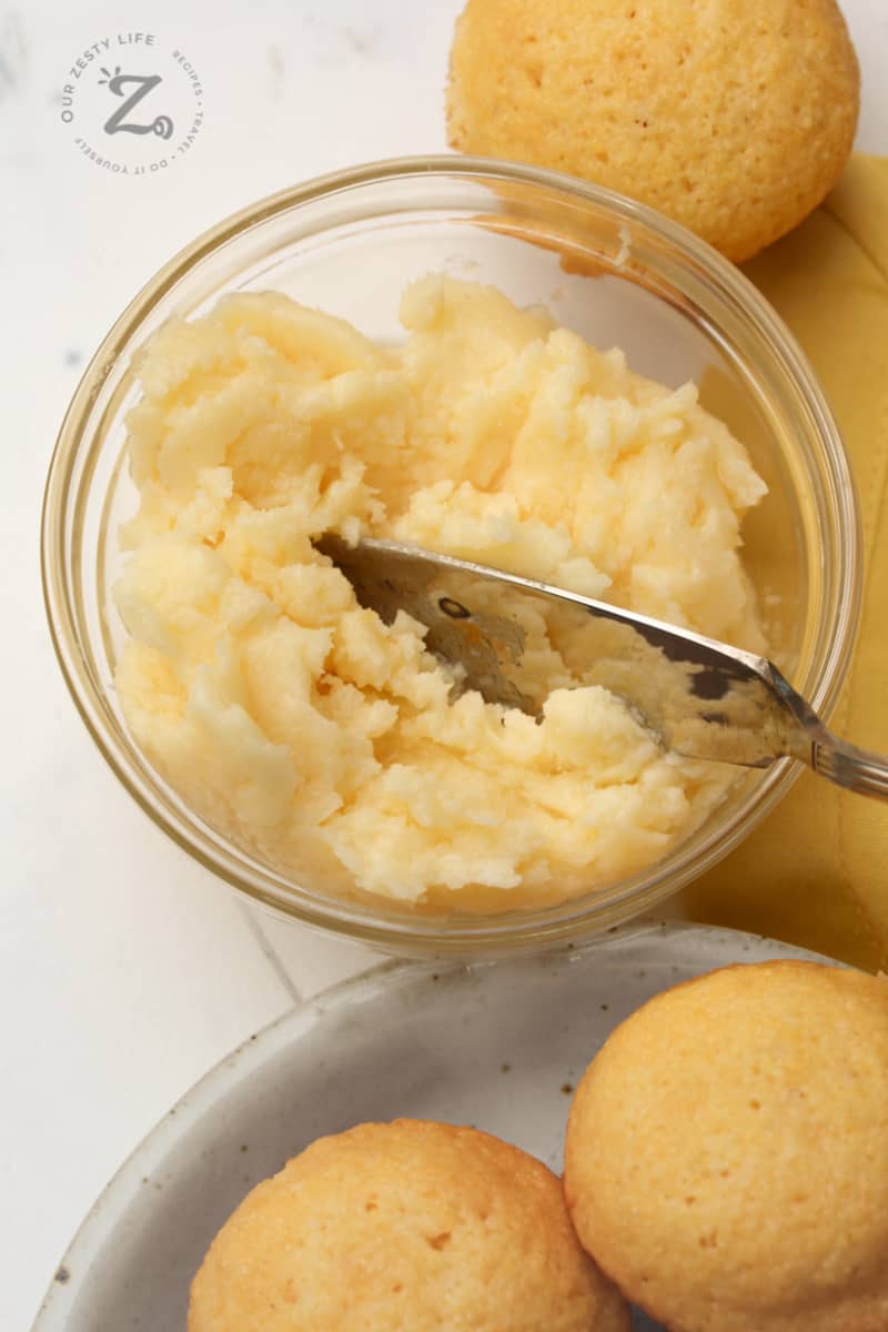 honey butter being in a glass bowl with a butter knife honey corn muffins on a plate and in the background
