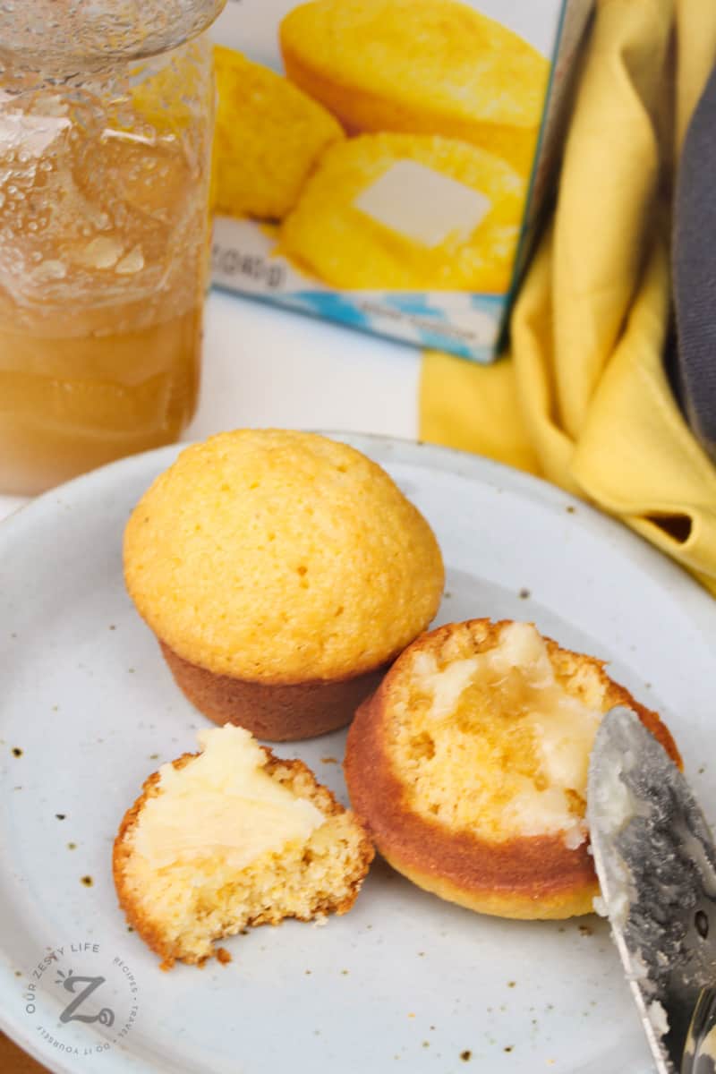 two honey cornbread muffins on a white plate, one broken open with honey butter on it, with a jar of honey and a box of Jiffy Corn Muffin Mix in the background