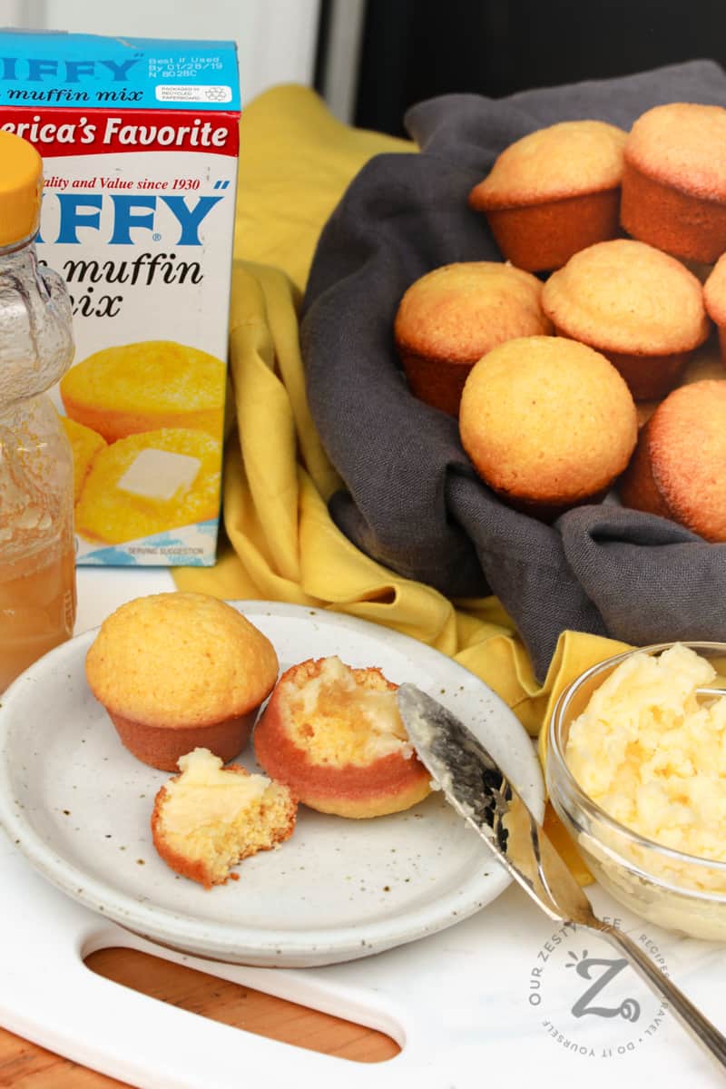 two honey cornbread muffins on a white plate, one broken open with honey butter on it, with honey butter with a knife, a jar of honey and a box of Jiffy Corn Muffin Mix in the background
