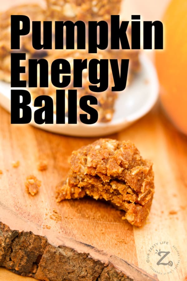 a pumpkin energy ball with a bite out of it on a wood board, with a white plate with pumpkin energy balls on it in the background