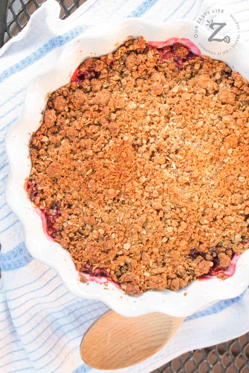 overhead of raspberry rhubarb crisp in a white pie dish with a wooden spoon and tea towel beside it