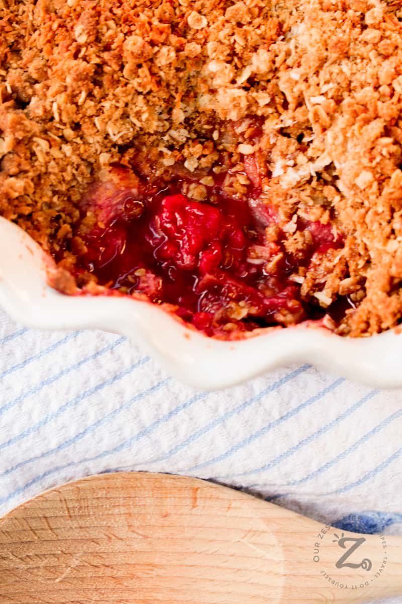 overhead closeup of raspberry rhubarb crisp in a white pie dish with a wooden spoon and tea towel beside it