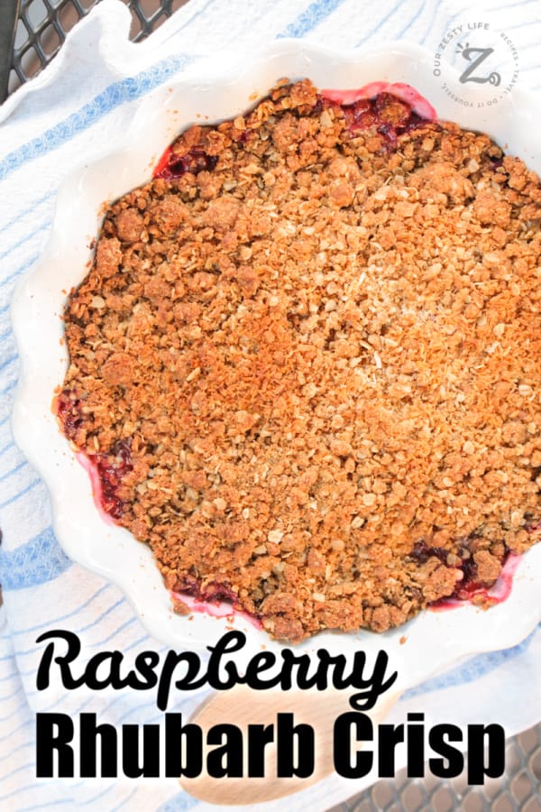 overhead of raspberry rhubarb crisp in a white pie dish with a wooden spoon and tea towel beside it