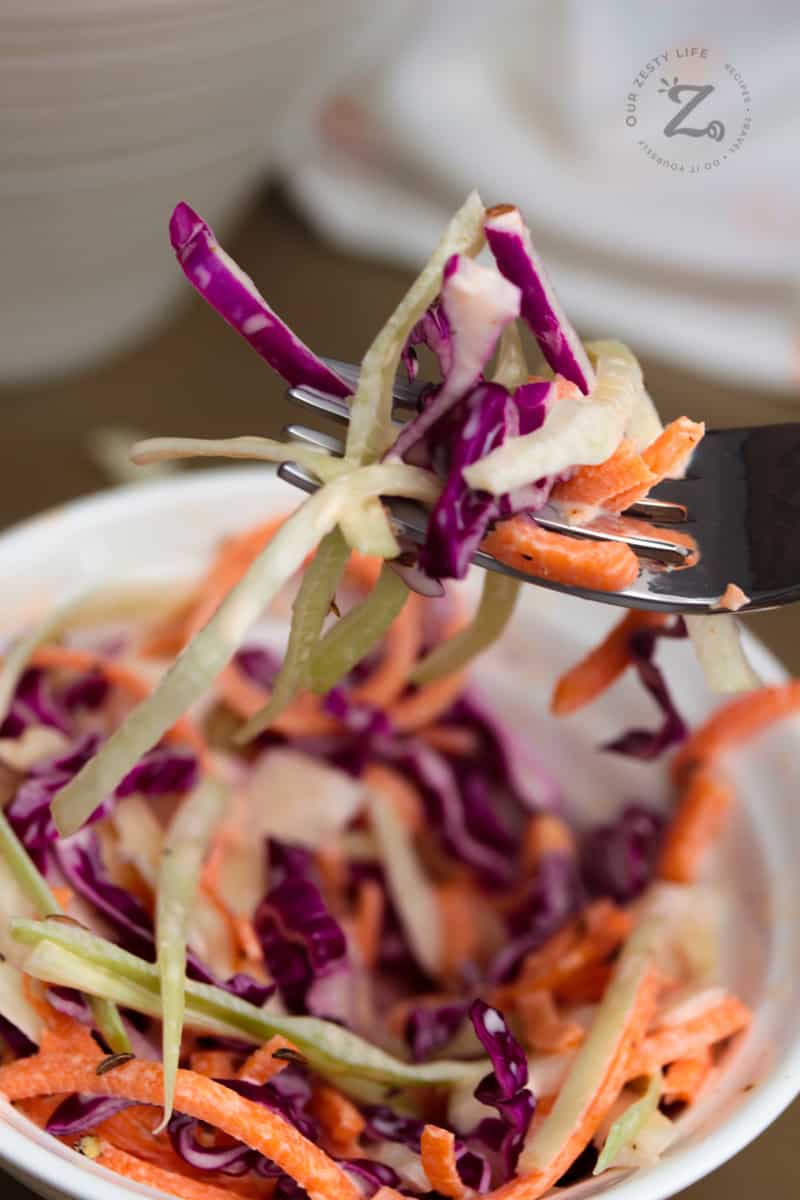 close up of creamy coleslaw with carrots and cabbage on a fork with a bowl of coleslaw in the background