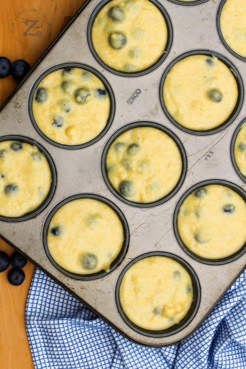 easy blueberry corn muffin batter in a muffin tin with blueberries and a blue checked towel in the background