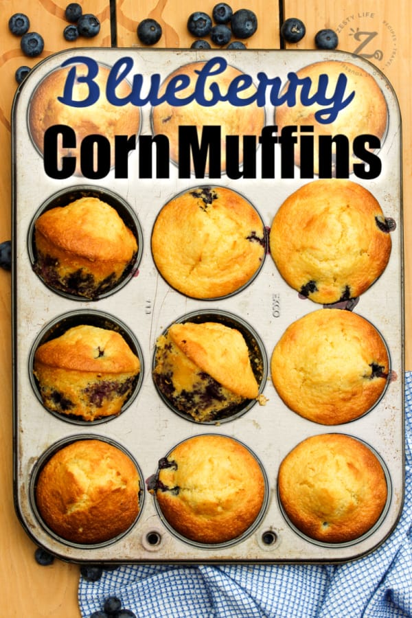 a muffin pan with 12 baked blueberry cornbread muffins, with blueberries, and a blue checked towel in the background