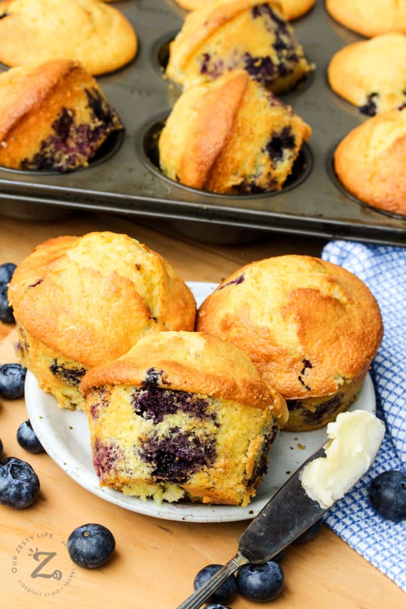 three blueberry corn muffins on a white plate with a butter knife slathered in butter, blueberries, a blue checked towel, and a pan of baked blueberry cornbread muffins in the background