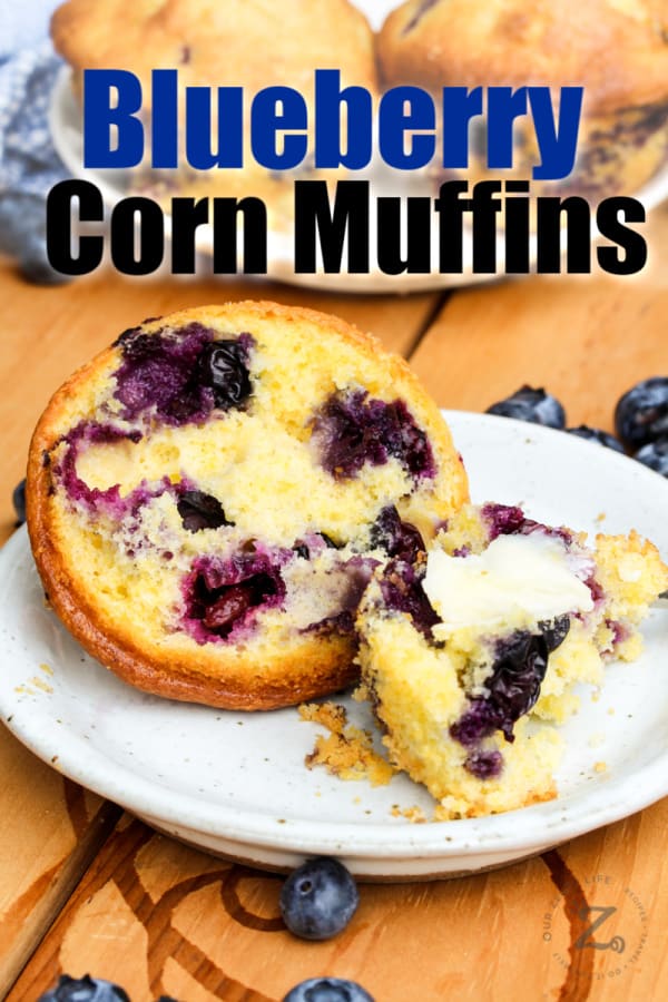a blueberry corn muffin open with butter on it on a white plate with blueberries and a plate with blueberry cornbread muffins in the background