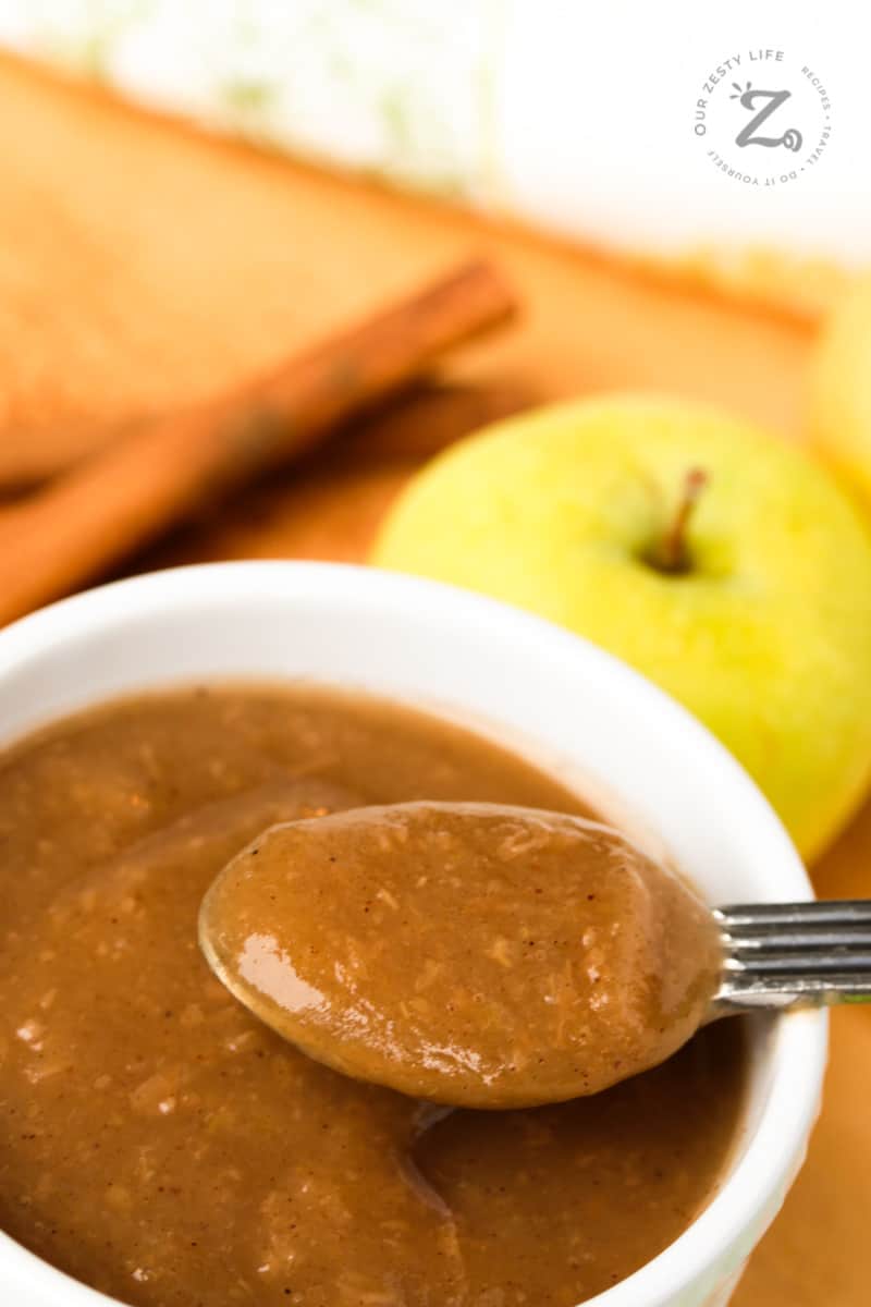 Instant Pot applesauce in a small white bowl with apples in the background and a spoon holding some applesauce in the foreground