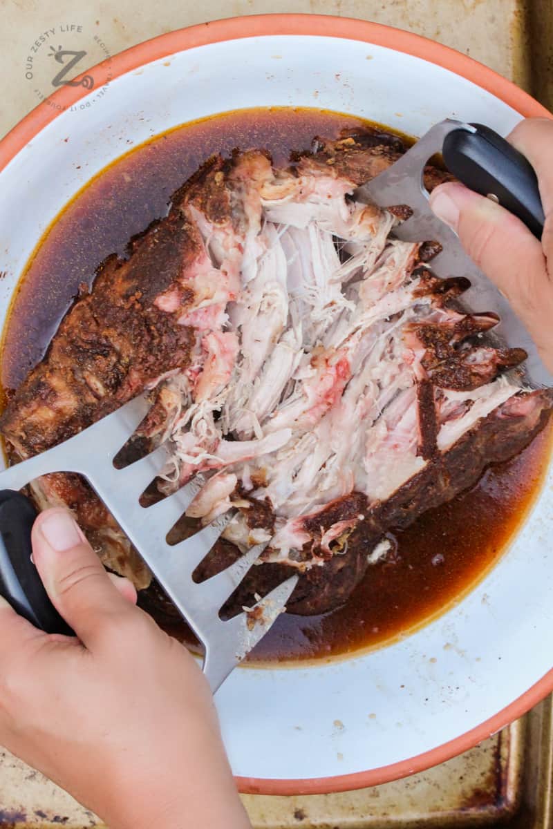 Shredding the finished Boston butt pork shoulder in a bowl with shredding tongs