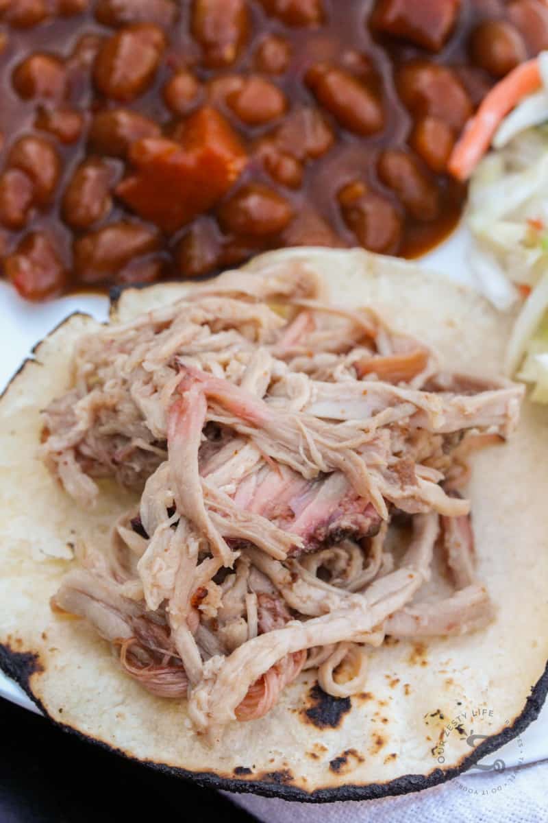 shredded pulled pork shoulder on a grilled corn tortilla, with baked beans and coleslaw in the background on the plate