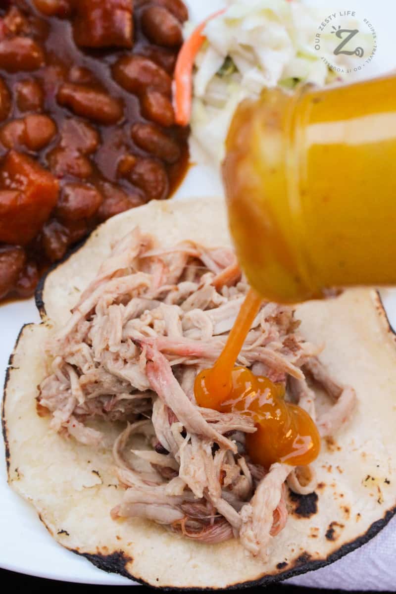 shredded pulled pork shoulder on a grilled corn tortilla, with added BBQ sauce, baked beans and coleslaw in the background on the plate