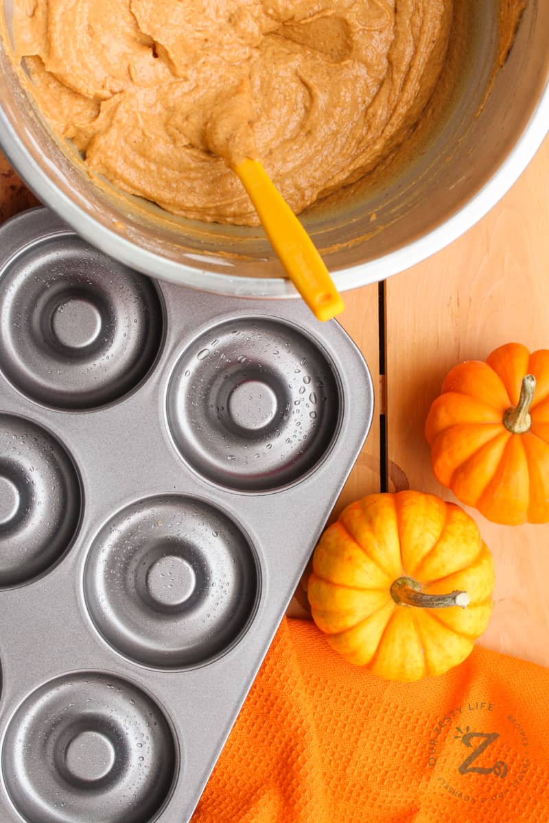 Pumpkin spice donut batter in a bowl and a donut baking dish beside it and 2 mini pumpkins beside the pan.