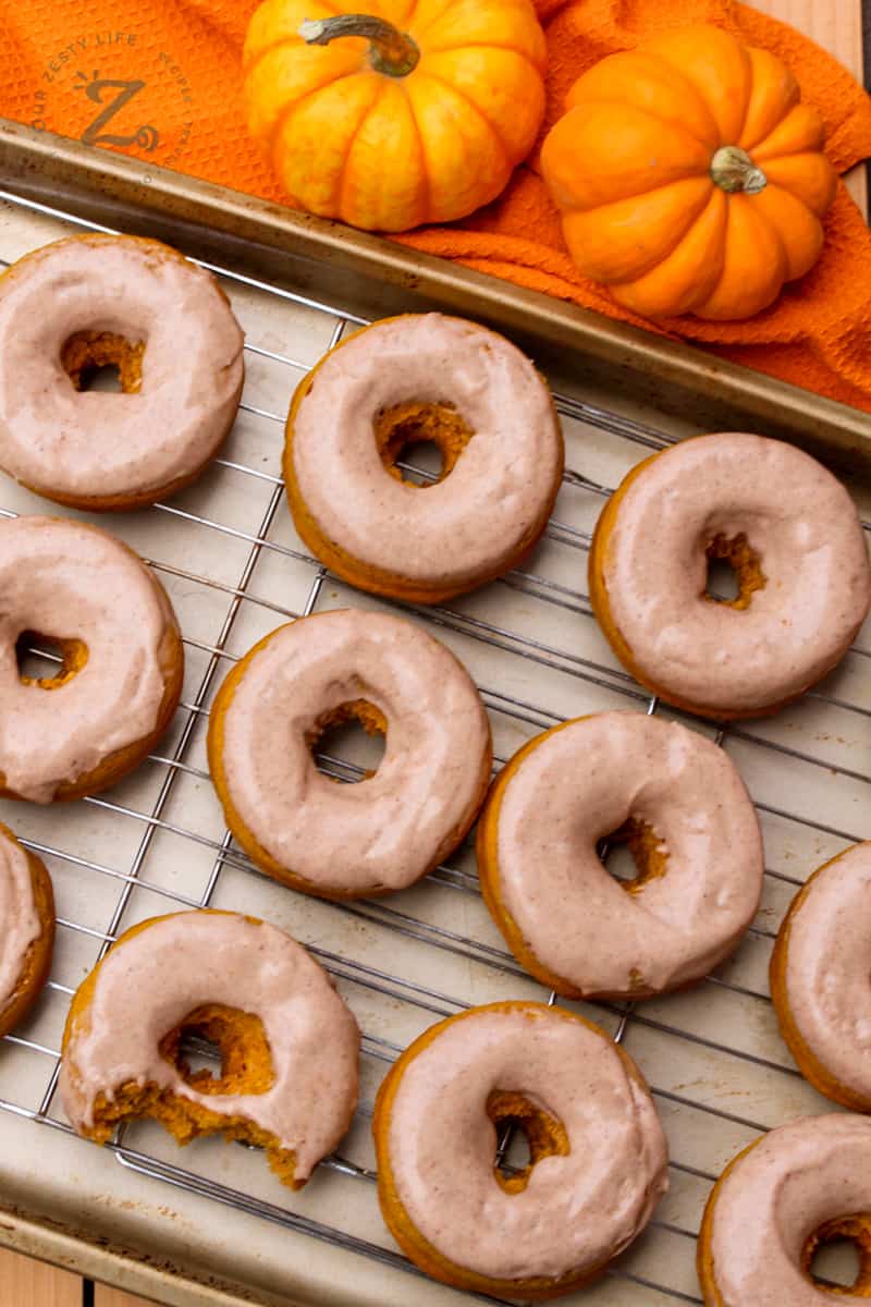 Pumpkin spice donuts with pumpkin spice glazed on a cooling rack and two mini pumpkins and an orange cloth in the background