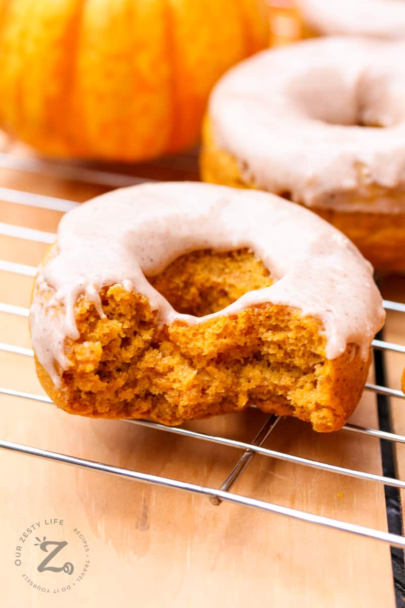Close up of a donuts in the foreground which has two bites taken out of it and the cooling rack has 1 mini pumpkin in it. 