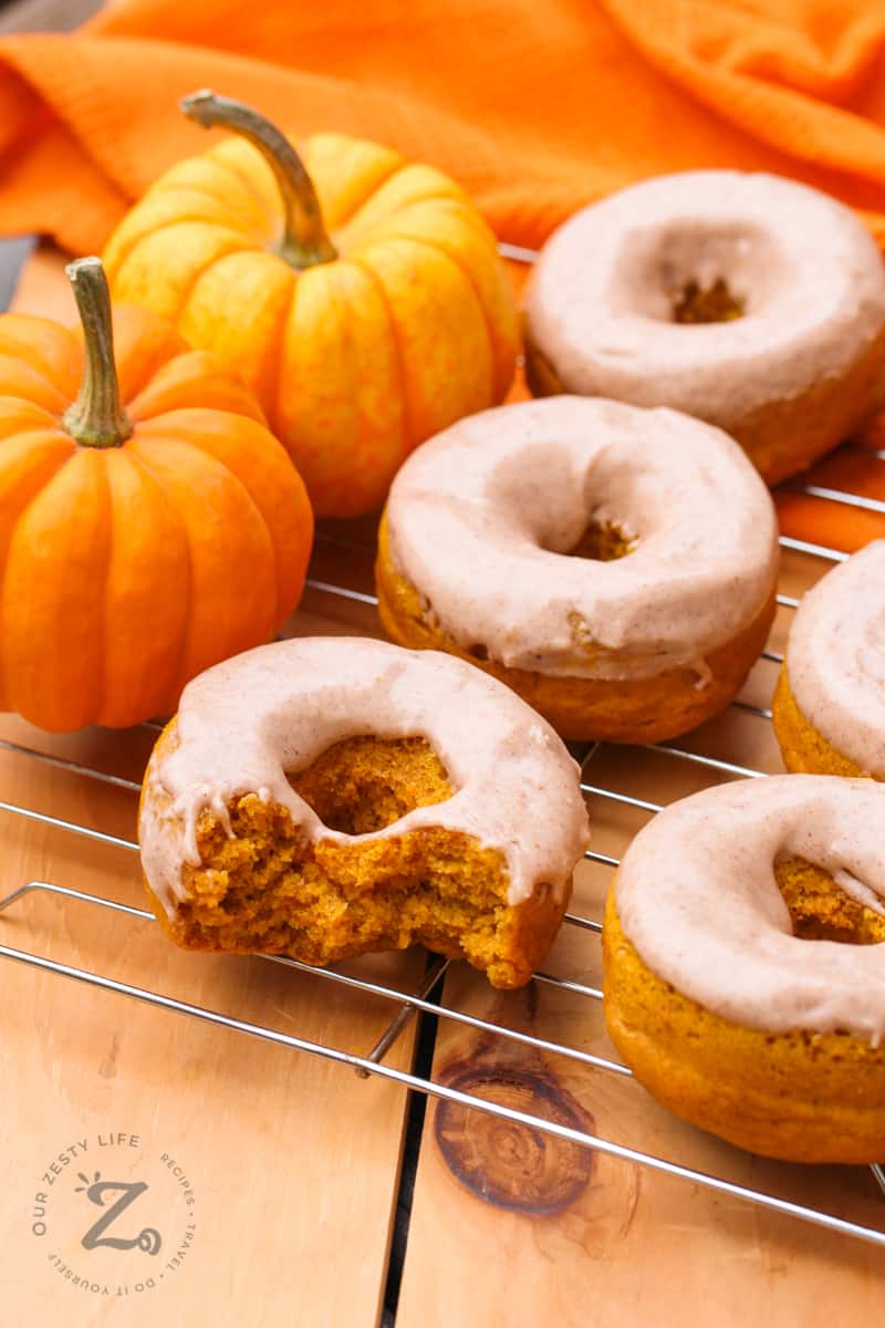 Baked pumpkin baked donuts with pumpkin spice glaze on a cooling rack. one of the donuts in the foreground has two bites taken out of it and the cooling rack with 2 mini pumpkins