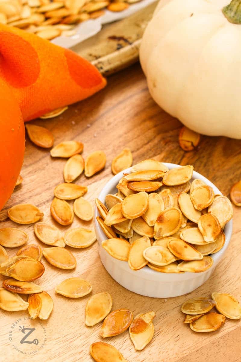 an overhead view of homemade roasted pumpkin seeds on a bowl overflowing onto a wooden platter and roasting pan with a white pumpkin and orange napkin in the background. 