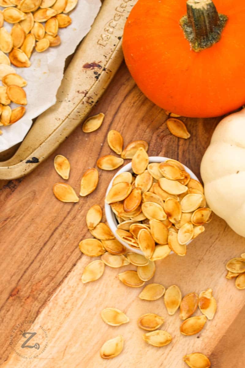 an overhead view of homemade roasted pumpkin seeds on a bowl overflowing onto a wooden platter and roasting pan with an orange and white pumpkin in the background. 
