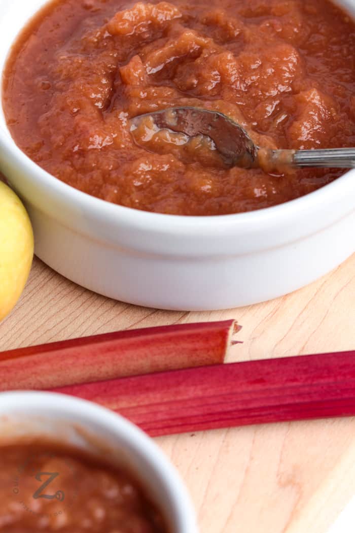 A close up of a bowl of Instant Pot rhubarb applesauce with a spoon stirring it and a couple of stalks of rhubarb in the foreground.
