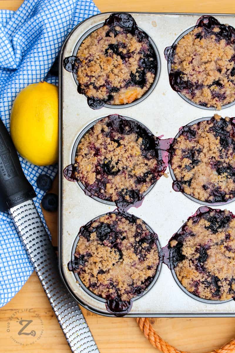 A tray of 6 cooked blueberry muffins in a pan and a lemon and zester in the background