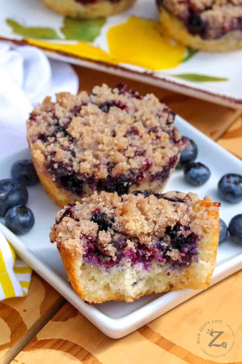 Blueberry muffins with crumb topping on a white plate - one of the muffins is cut in half so you can see the inside