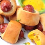 close up of 5 mini pigs in a blanket beside some mustard dipping sauce one has a bite taken from it