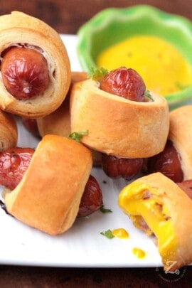 close up of 5 mini pigs in a blanket beside some mustard dipping sauce one has a bite taken from it