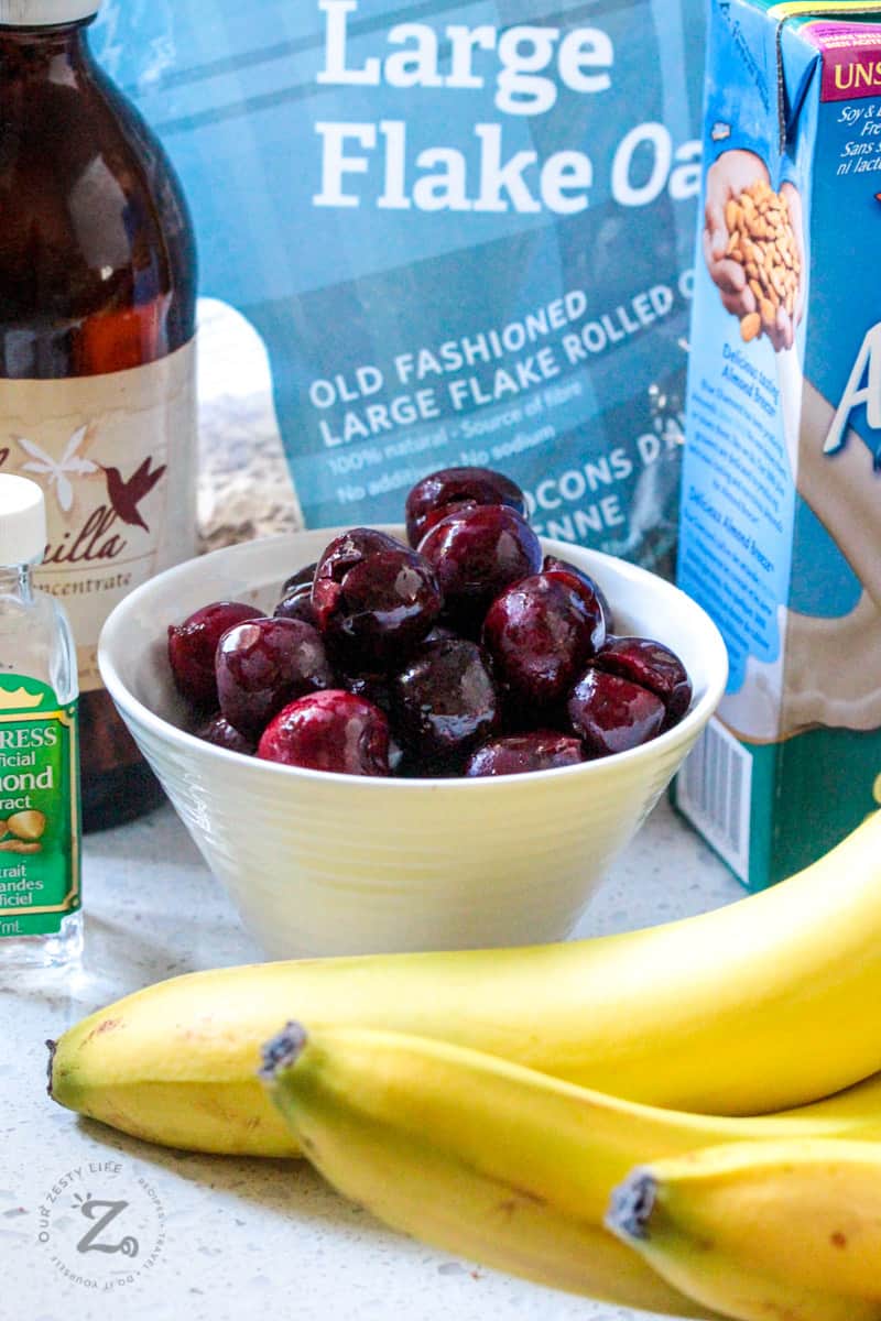 side view of cherries in a white bowl with bananas in the foreground and packages of almond milk and large flake oats in the background