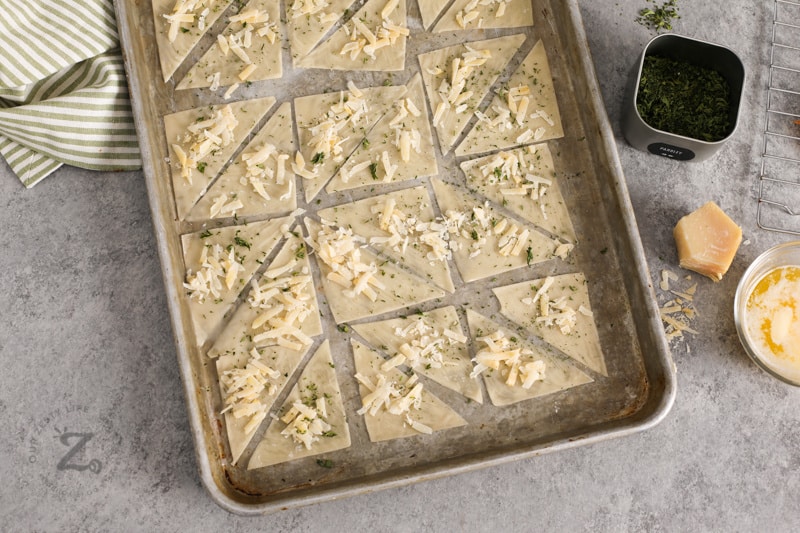 Overhead of uncooked parmesan and parsley wonton crackers laid out on a cookie sheet.