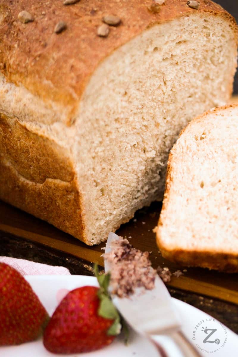 A loaf of whole wheat bread on a cutting board with a slice cut out of it and some strawberries on plate with strawberry butter on a knife in the foreground.