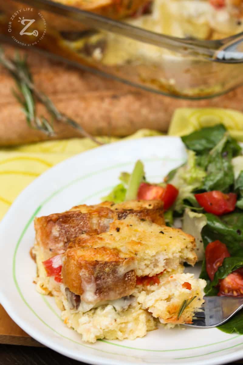 A served slice of breakfast strata with sausage on a plate with a green salad and the pan of strata in the background