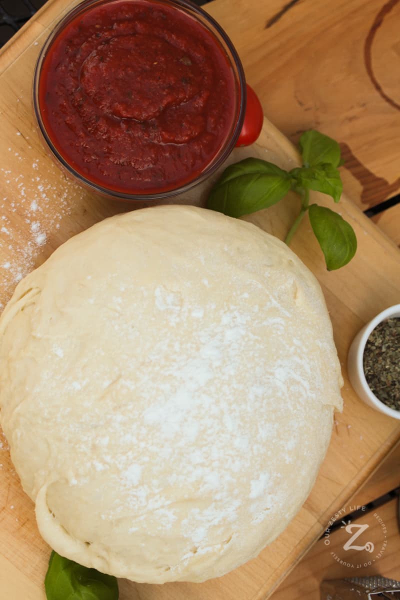 A ball of homemade pizza dough on a rolling board sprinkled with flour with some basil and tomato sauce on the side