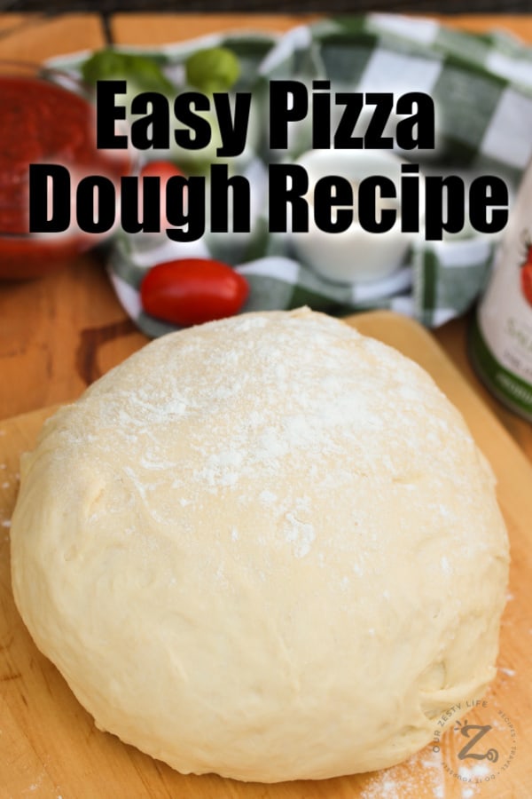 A ball of homemade pizza dough on a rolling board sprinkled with flour with pizza ingredients in the background
