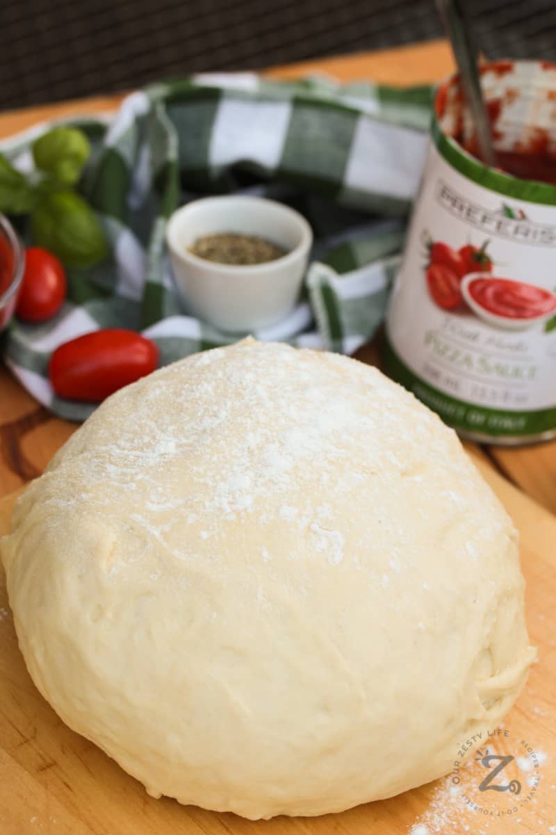 A ball of homemade pizza dough on a rolling board sprinkled with flour with pizza ingredients in the background