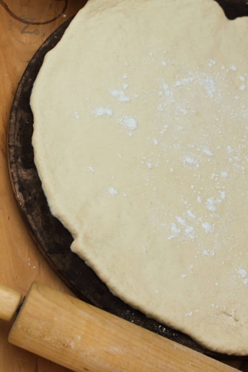 Homemade pizza dough rolled out on a pizza stone, uncooked with a rolling pin in the foreground